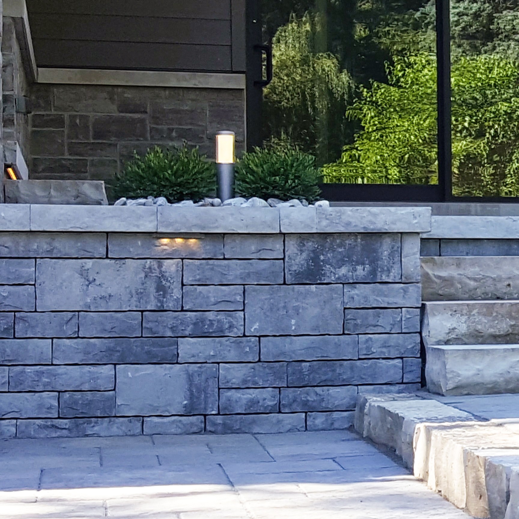 Stone steps and wall in front of a modern building entrance with a reflective glass door and surrounding greenery.