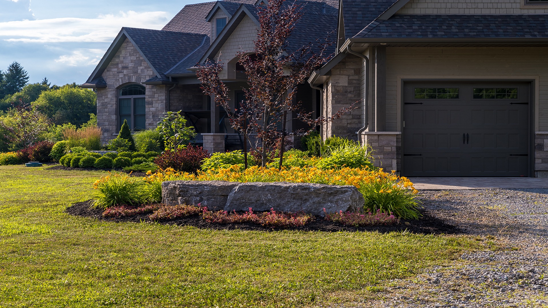 A stone house with lush landscaping, featuring colorful flowers, shrubs, and a small tree. Nearby, a stone path and neatly trimmed lawn.