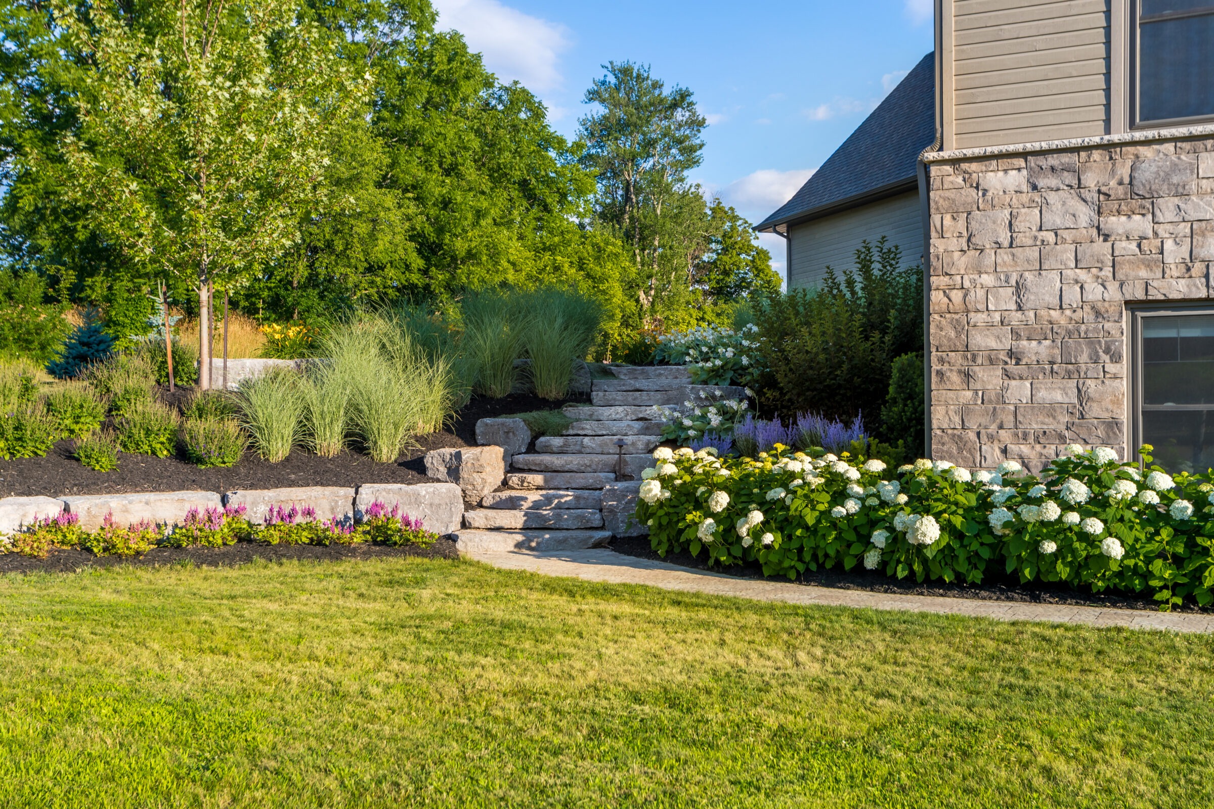 A beautifully landscaped garden with stone steps, vibrant flowers, and lush greenery adjacent to a stone-walled house under a clear blue sky.