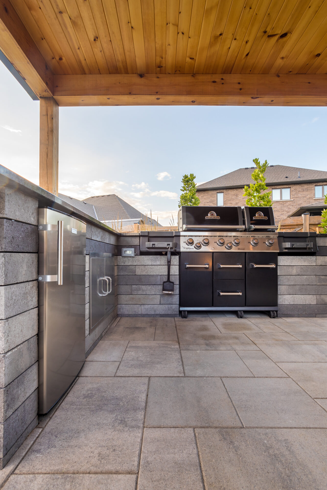 Outdoor kitchen setup with grill, refrigerator, and stone counter. Residential houses in the background, under a wooden pergola roof.
