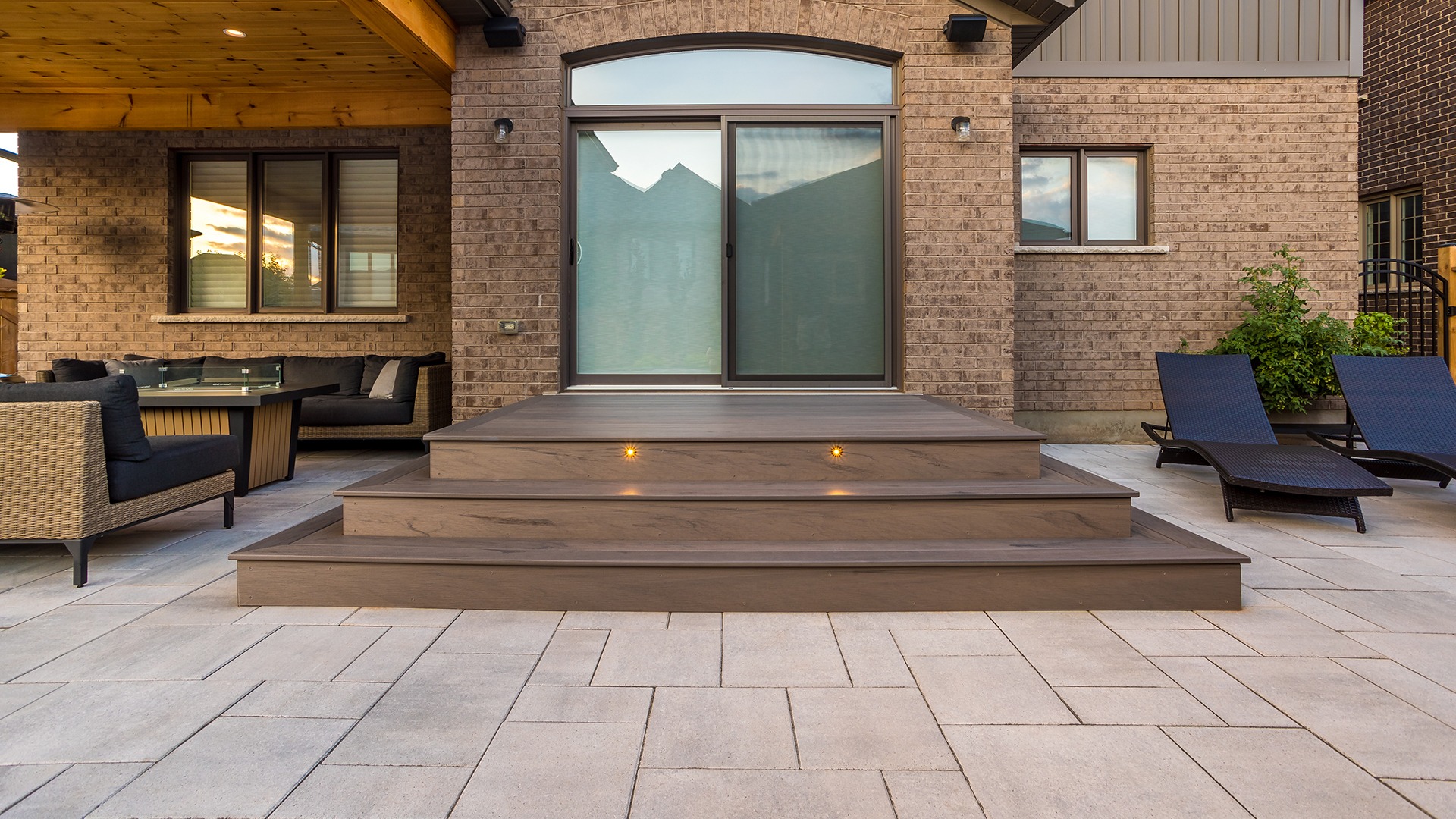 Modern patio with beige bricks, wooden deck, lounge chairs, and black wicker furniture. Steps have built-in lights; glass door and large windows.