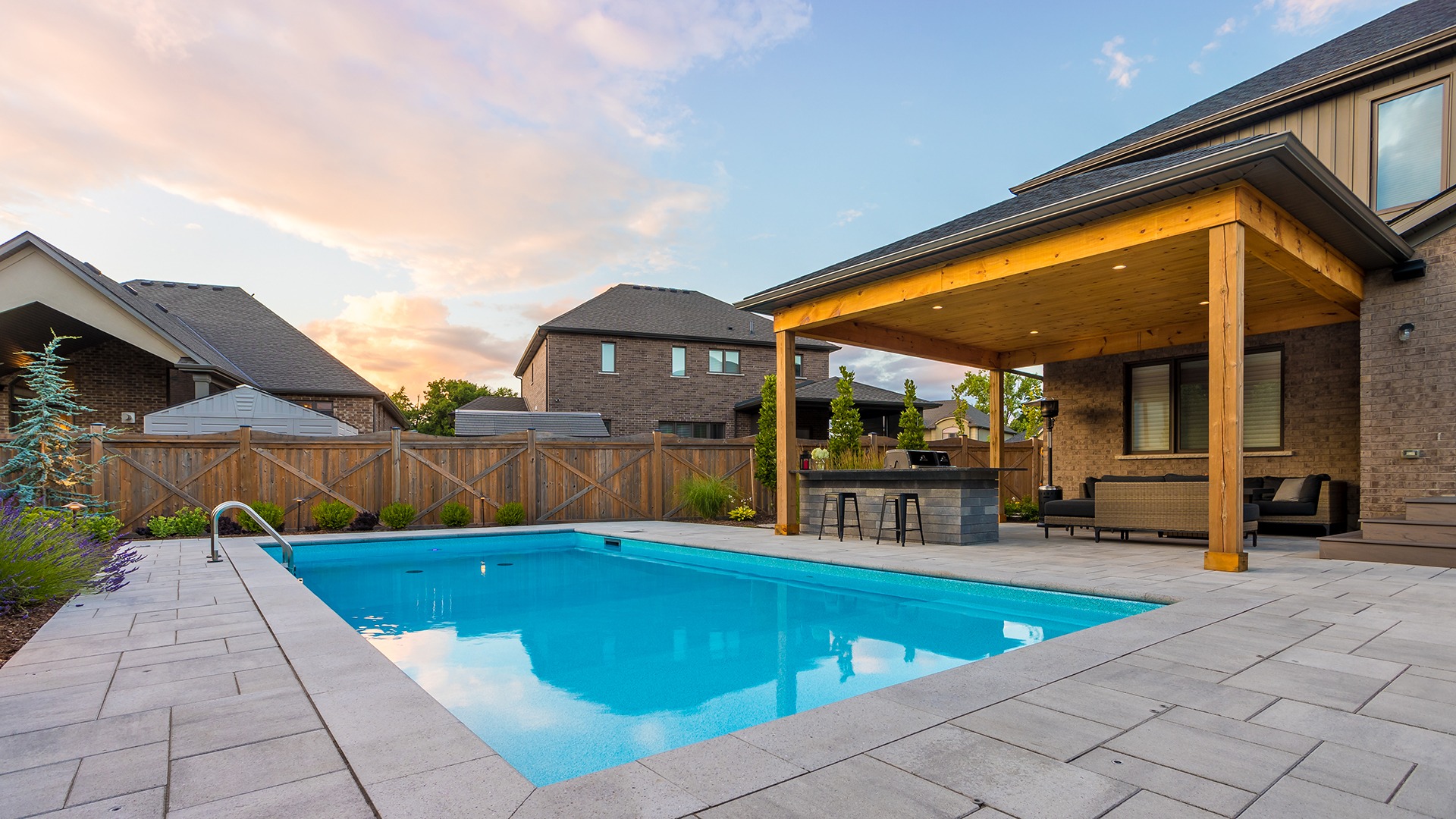 Modern backyard with a swimming pool, wooden pergola, and seating area. Surrounded by brick houses and wooden fences. Clear sky at sunset.