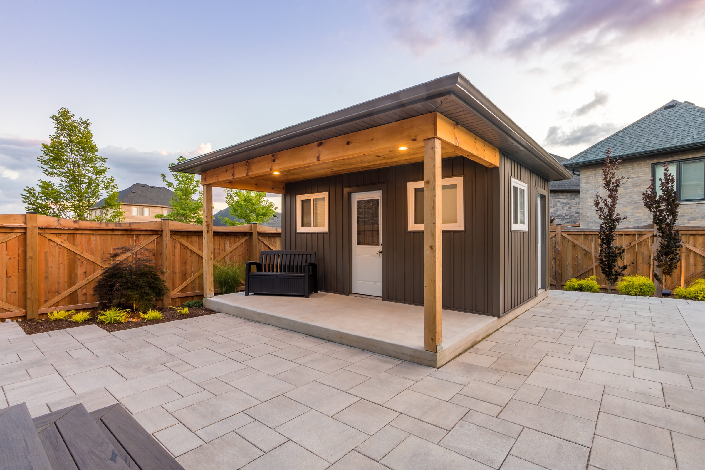 A modern backyard features a small shed with a covered porch, surrounded by a wooden fence and tidy landscaping under a cloudy sky.