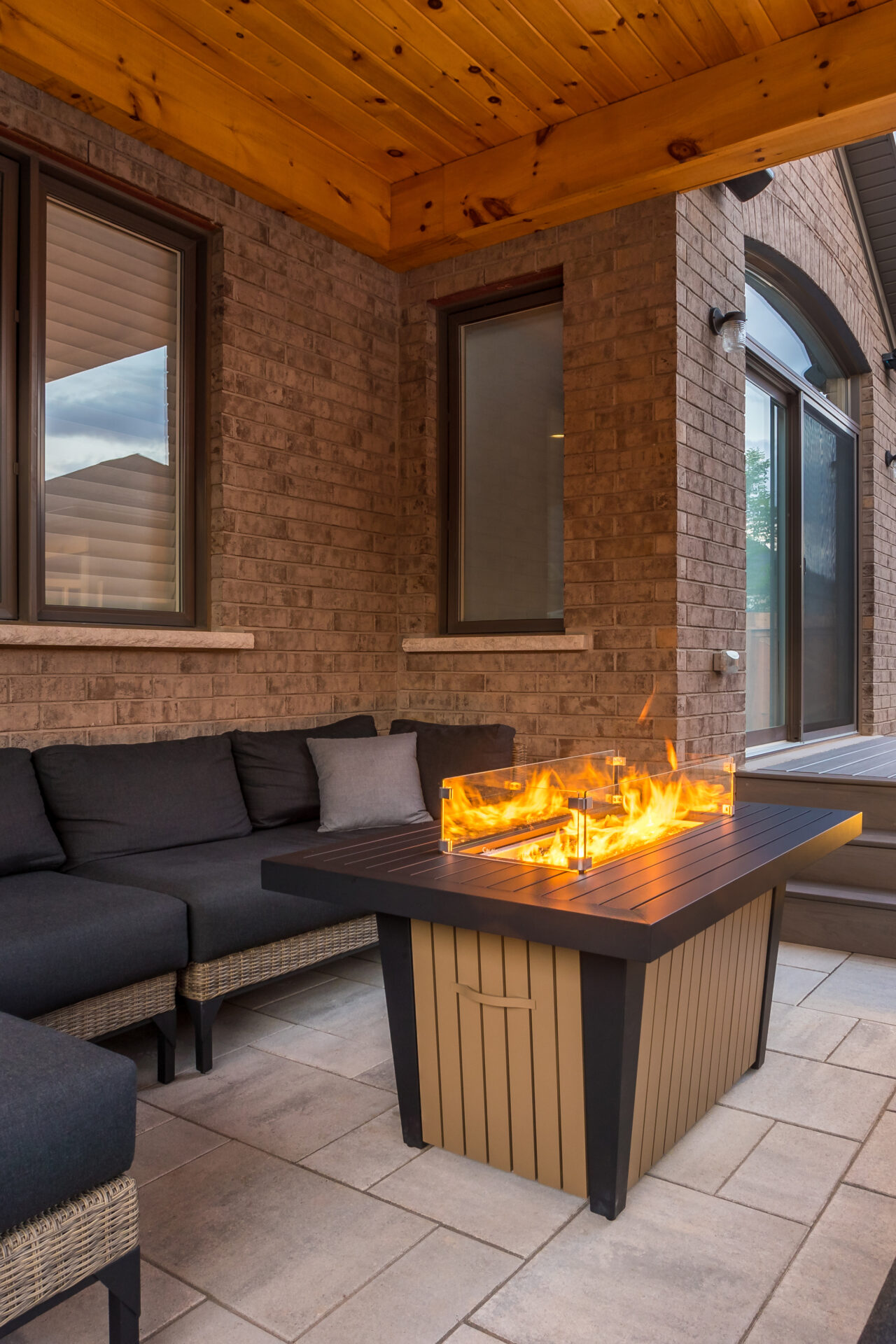 Cozy patio with a modern fire pit, surrounded by cushioned seating. The warm atmosphere is accentuated by brick walls and wooden ceiling.