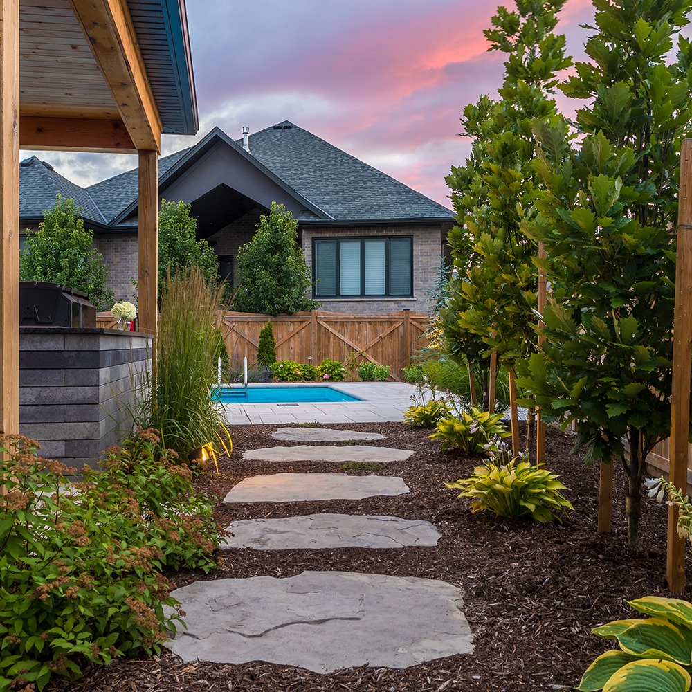 Stone path leads to a pool in a backyard garden, framed by lush greenery and a wooden fence, under a colorful sunset sky.