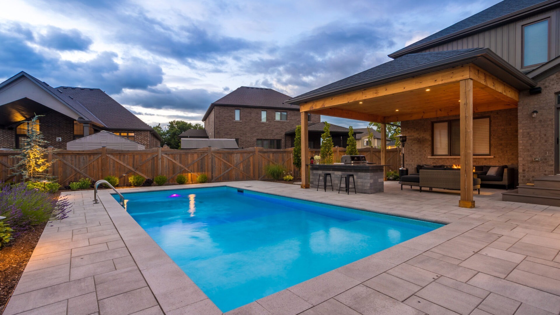 A backyard features a modern pool, cozy seating under a wooden pergola, and adjacent brick house, surrounded by a wooden fence and garden.