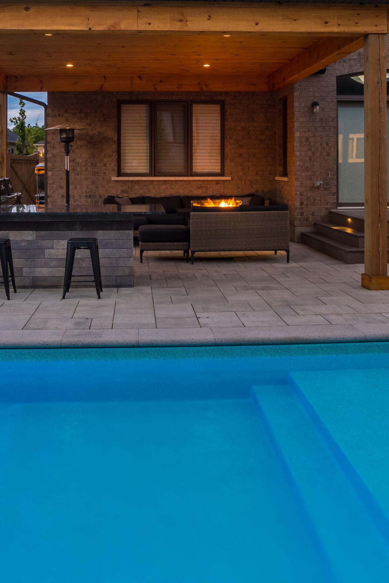 A modern backyard features a swimming pool, patio seating area, fire pit, and covered wooden pergola, creating an inviting outdoor retreat at twilight.
