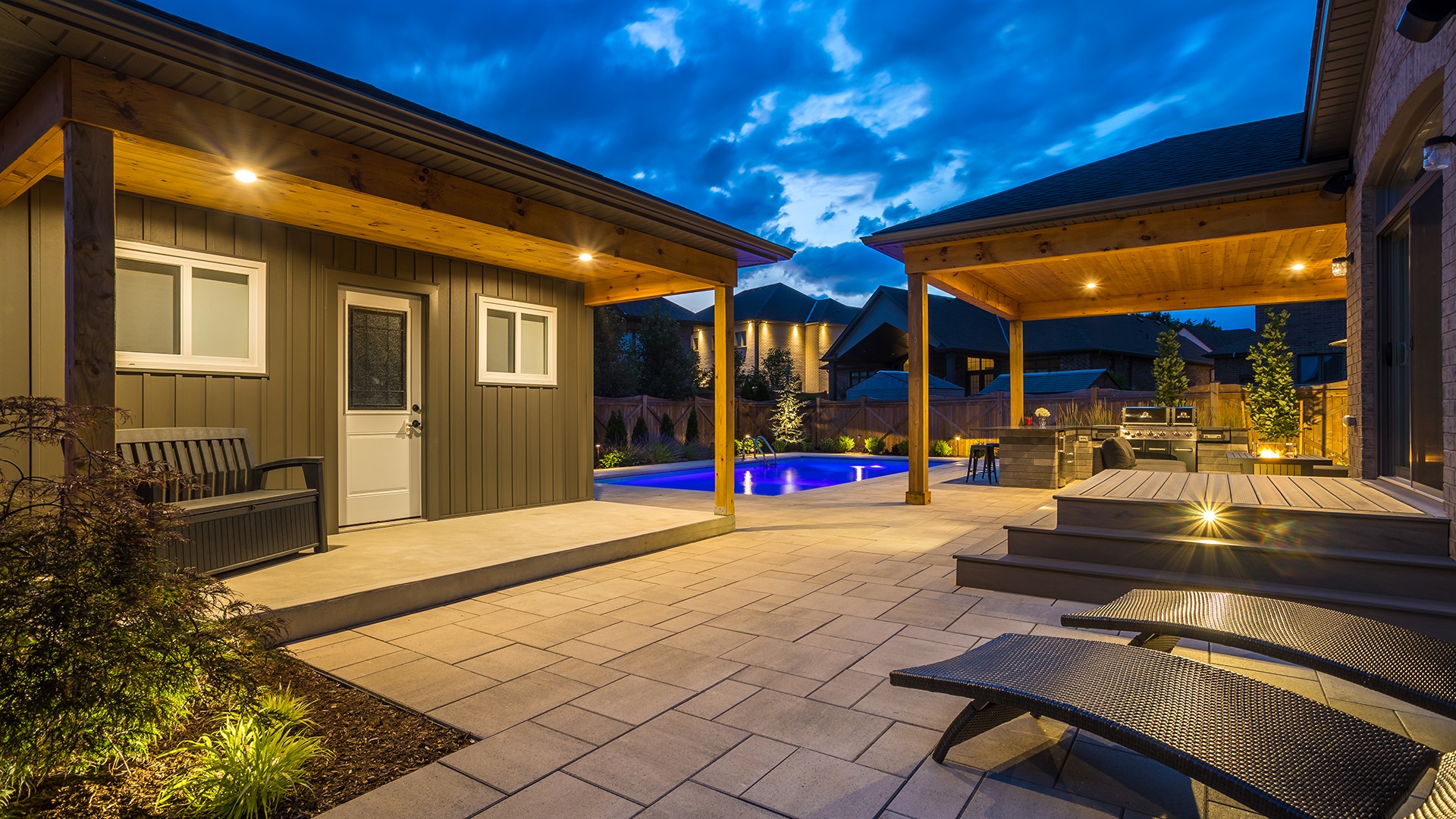 Modern backyard with illuminated pool, patio, and pergola at dusk. Features outdoor kitchen, seating, and ambient lighting. Quiet residential setting with no people.
