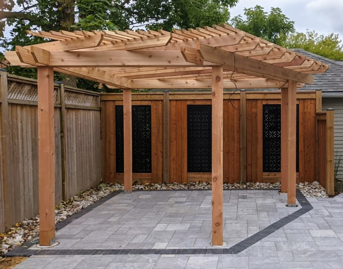 A wooden pergola stands on a neatly paved patio, enclosed by a wooden fence, surrounded by greenery on a cloudy day.