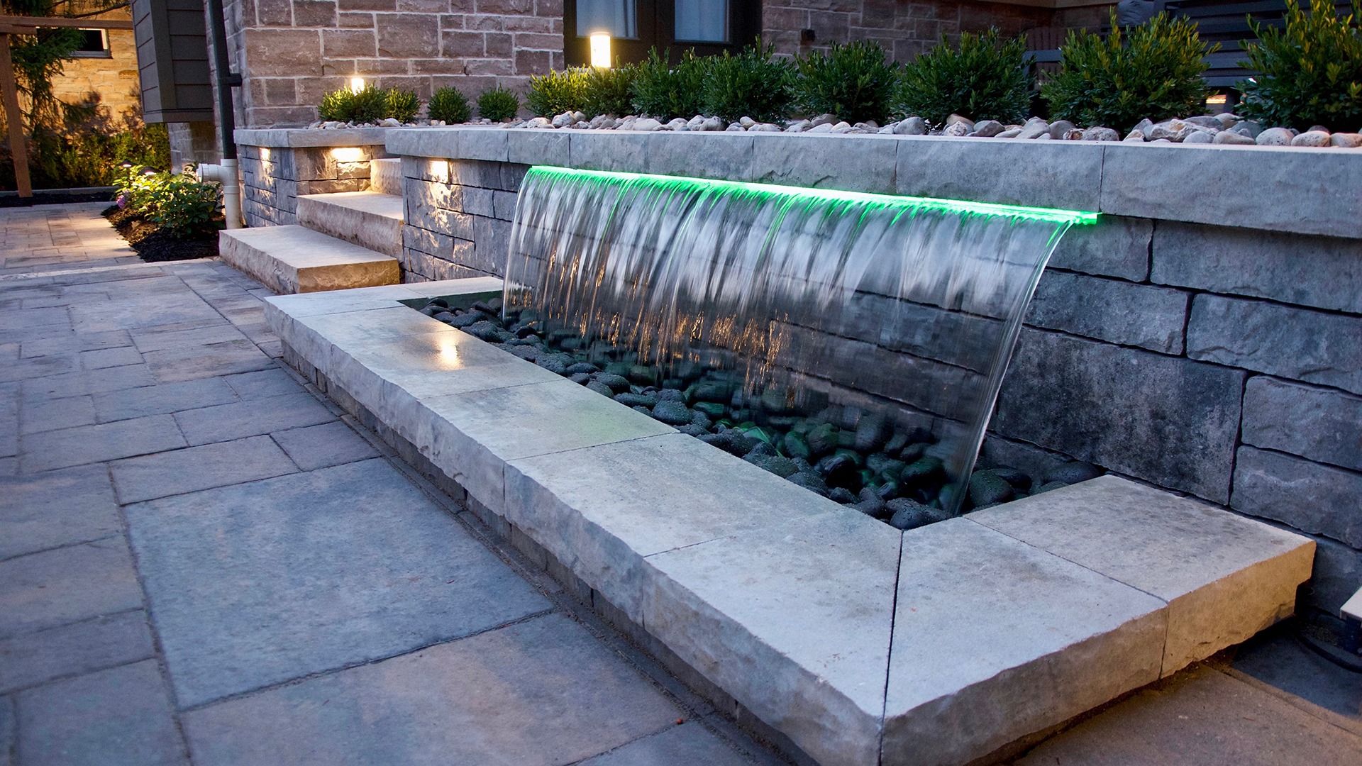 Modern patio with stone waterfall feature, illuminated by green light, surrounded by plants. Evening atmosphere with textured stone elements.