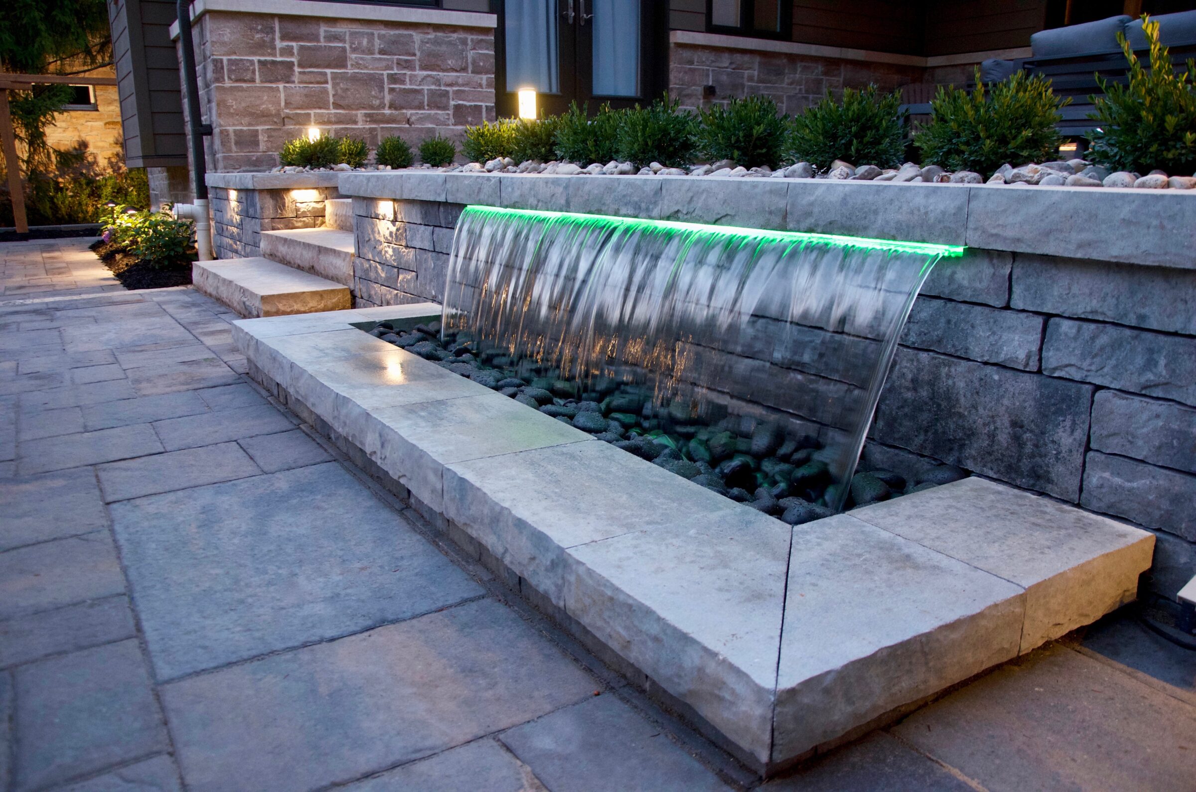 Modern outdoor stone patio featuring a sleek waterfall, illuminated with green lights, surrounded by neatly arranged plants and textured brick walls.