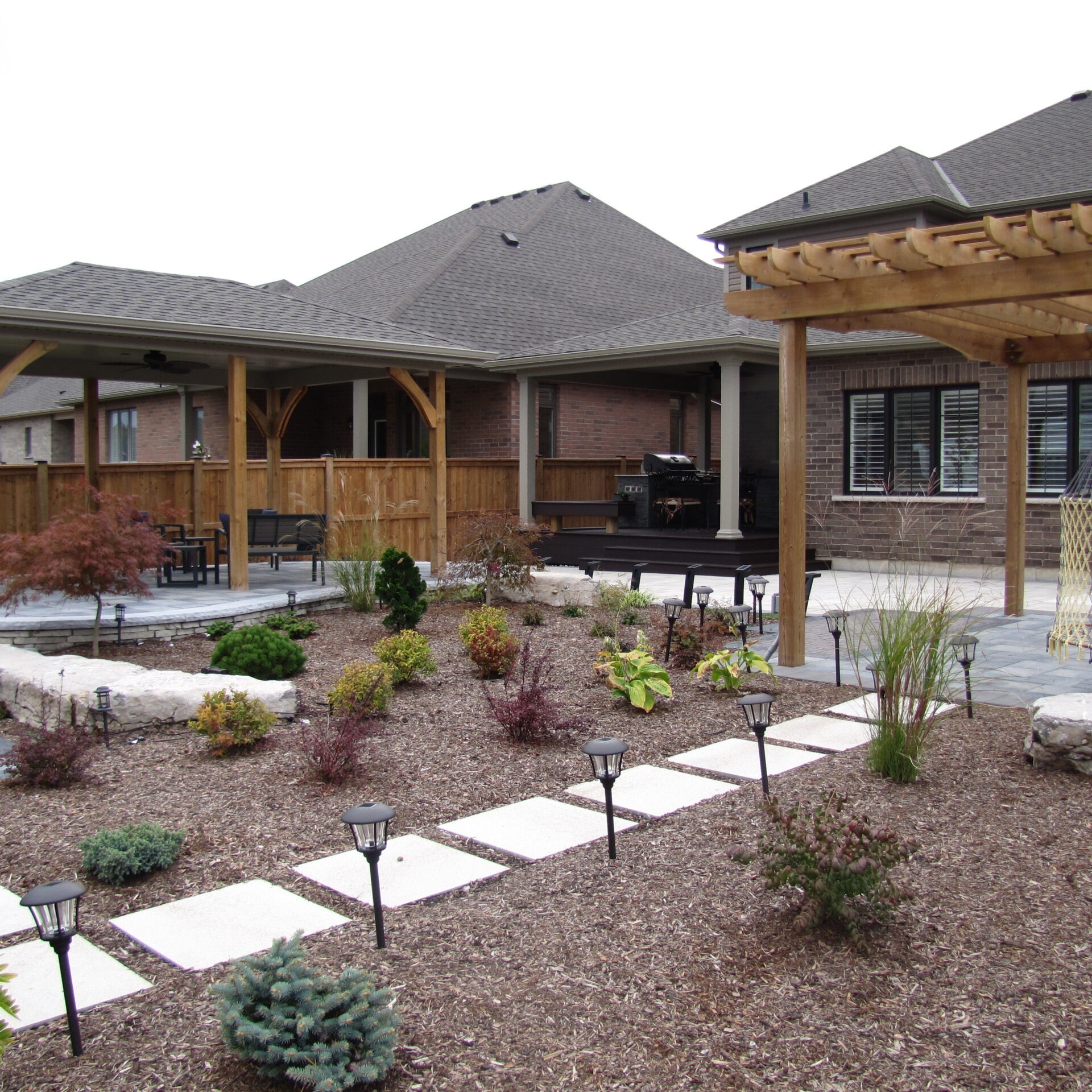 A landscaped backyard features a wooden pergola, patio furniture, neatly arranged plants, pathway lights, and an outdoor grill area under a covered structure.