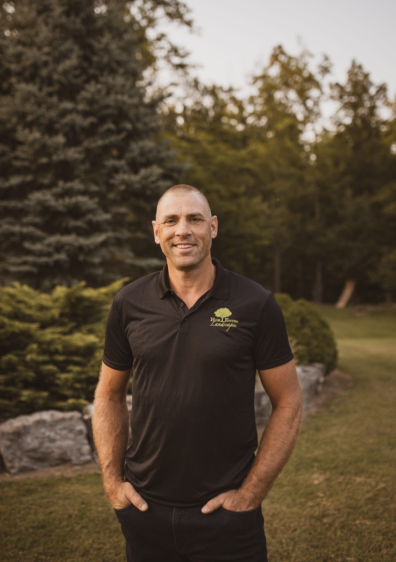 A person in a black shirt stands outdoors, surrounded by trees and rocks, smiling contently on a lush green lawn.