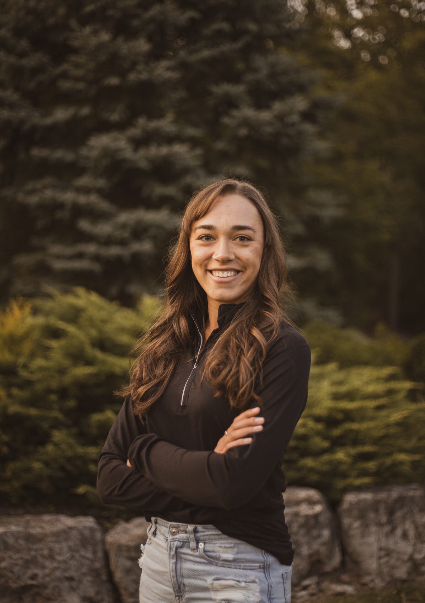 A person with long brown hair and a black sweater smiles, standing outdoors with a tree and greenery in the background.