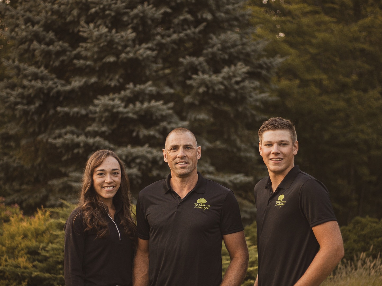 Three people in matching dark shirts stand outdoors, surrounded by lush greenery and trees, smiling at the camera.