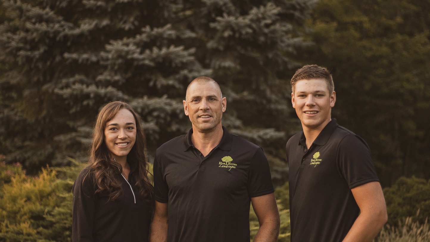 Three people wearing black polos standing outdoors, smiling, with lush evergreen trees in the background, creating a warm and friendly atmosphere.