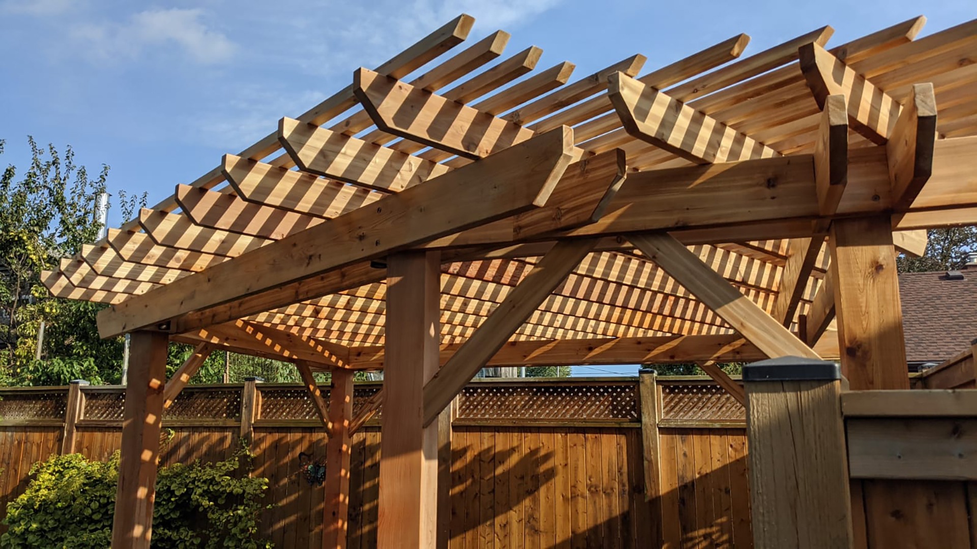 A wooden pergola casts striped shadows on a patio. Sunny day with blue sky, surrounded by a wooden fence and lush greenery.