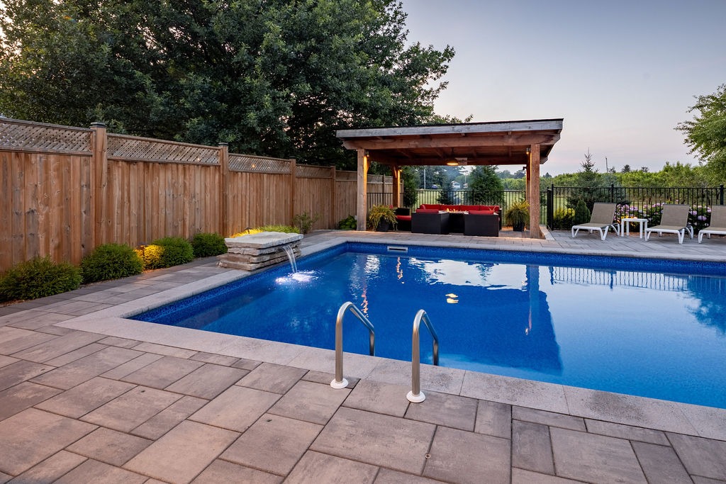 A backyard features a swimming pool, cozy seating under a wooden pergola, surrounded by a wooden fence, plants, and loungers against scenic greenery.