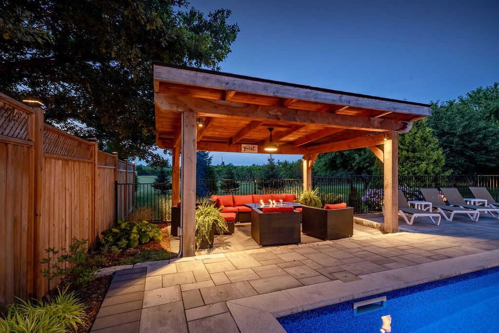 A cozy outdoor seating area with red cushions under a wooden pergola beside a pool, surrounded by lush green trees and fence.