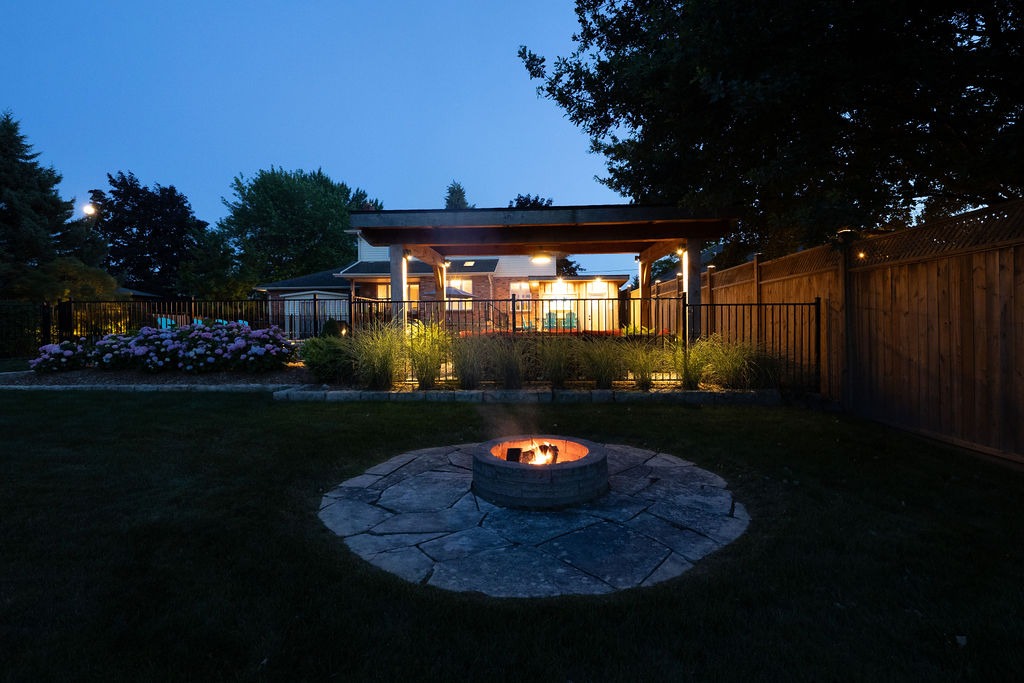 A peaceful backyard at dusk features a fire pit, patio with warm lighting, and lush greenery, enclosed by a wooden fence.