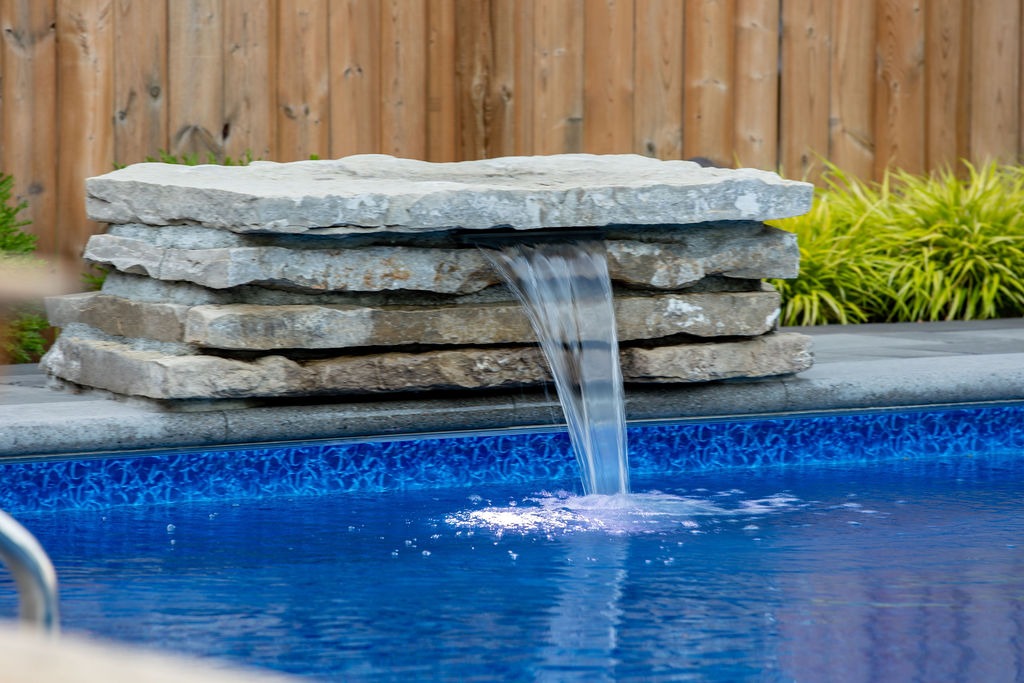 A small waterfall feature flows into a blue swimming pool surrounded by plants and a wooden fence, creating a tranquil garden scene.