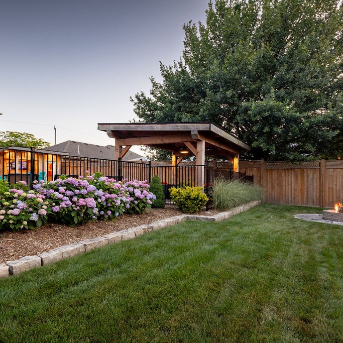 A backyard garden with blooming flowers, a wooden pergola, and a small fire pit, surrounded by a wooden fence and trees.