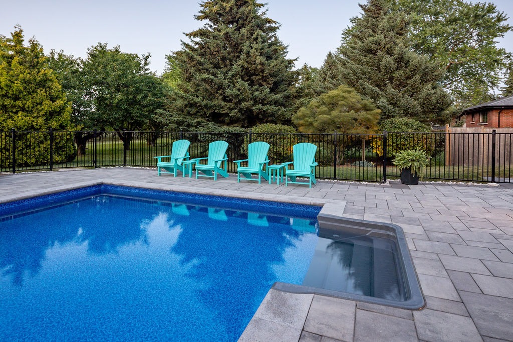 A tranquil backyard features a clear blue pool, surrounded by lush greenery and four blue chairs under a serene sky, enclosed by a fence.