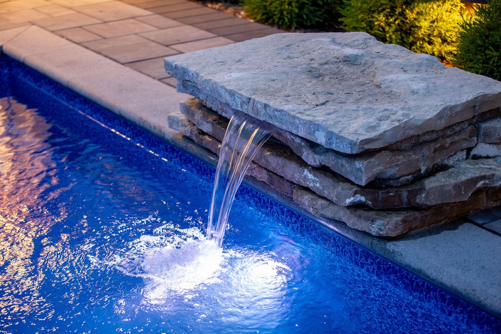 A serene poolside scene with water cascading from a stone fountain under soft evening lights, surrounded by paving stones and greenery.