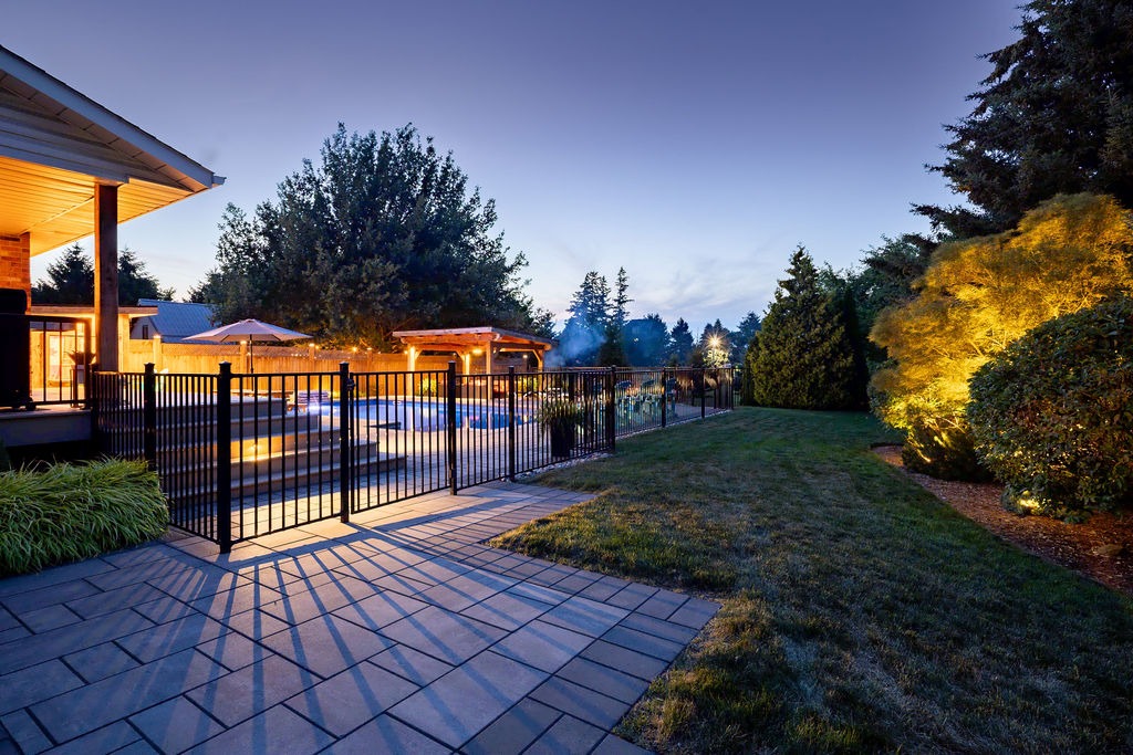 A modern backyard features a lit swimming pool surrounded by a fence, trees, and patio areas, with soft evening lighting enhancing the atmosphere.