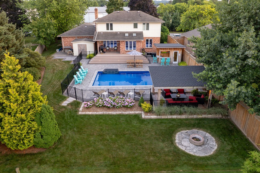 A backyard features a pool, deck, seating area, and lush landscaping, adjacent to a two-story brick house surrounded by trees and greenery.