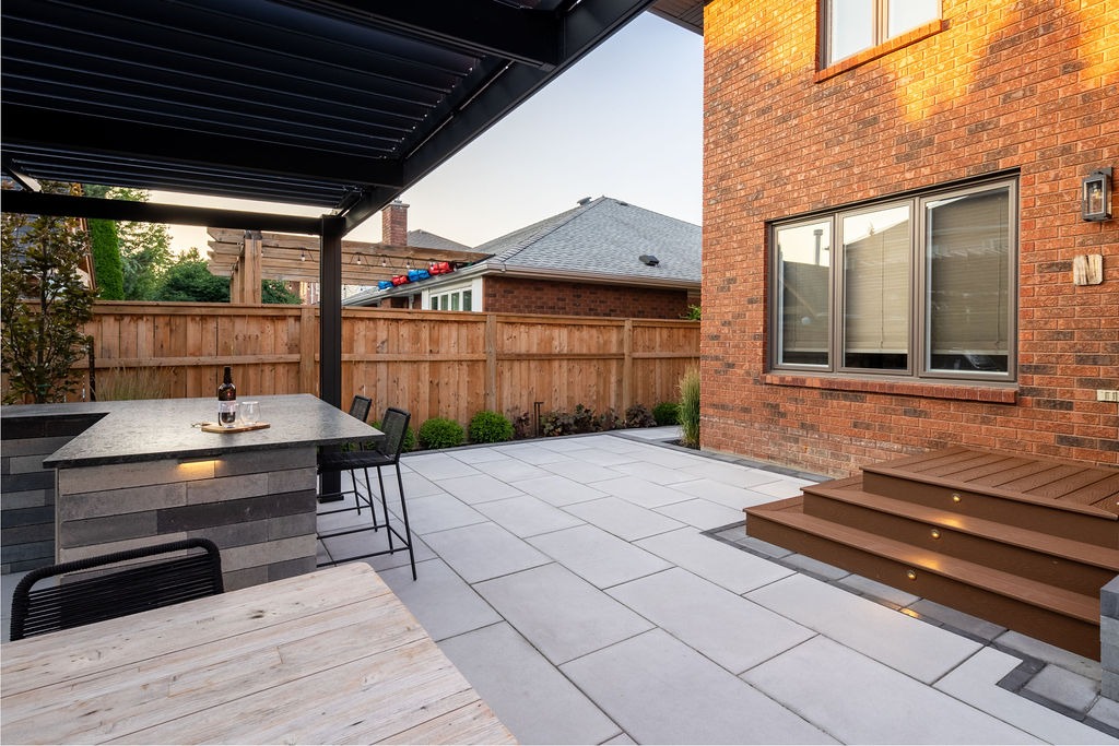 Modern backyard patio features seating area, brick house, and wooden fence. Includes outdoor furniture with bottle on table. No recognizable landmarks or people.