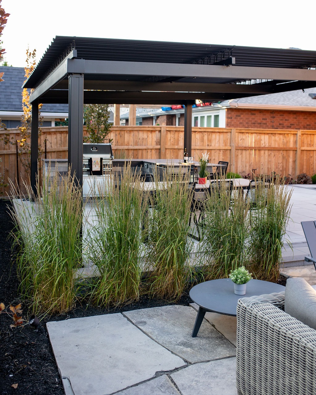 Modern backyard patio with pergola, outdoor furniture, and a grill. Tall grasses and wooden fencing provide privacy in a cozy outdoor setting.