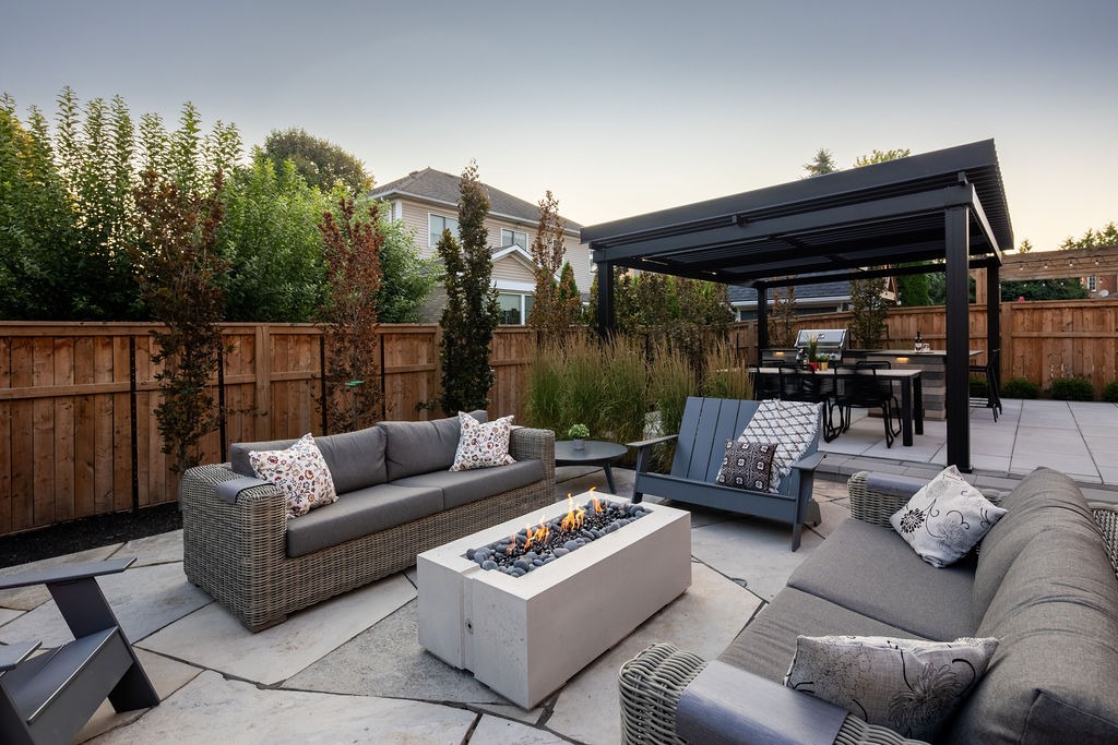 Modern backyard with cozy seating, fire pit, and pergola-covered dining area, surrounded by wooden fencing and lush greenery, creating a warm, inviting atmosphere.