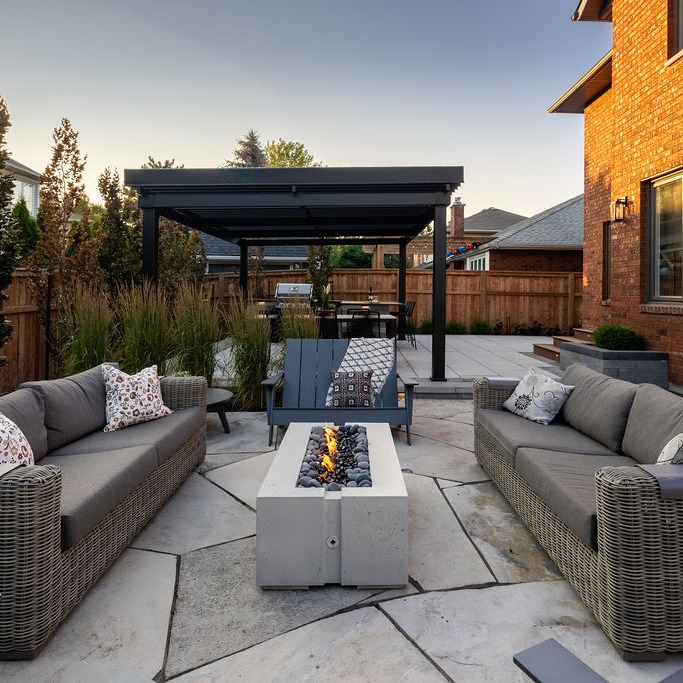 Modern backyard patio with pergola, fire pit, wicker sofas, and decorative pillows. Surrounded by wooden fence, vibrant plants, and brick house.