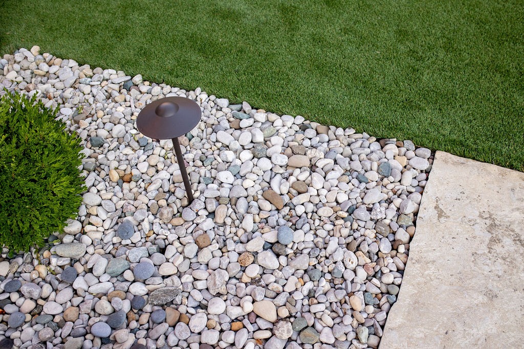A landscaped garden features smooth rocks, a metal garden light, lush greenery, and a neatly cut grass patch alongside a stone pathway.