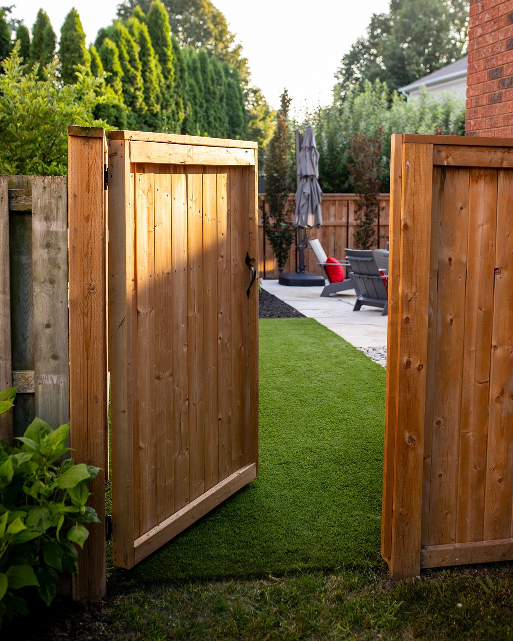 Open wooden gate leads to a patio with chairs, umbrella, and lush greenery under soft, evening light. Cozy backyard setting.