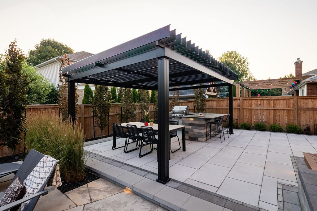Modern backyard patio with a pergola, dining area, and built-in grill. Surrounded by a wooden fence and landscaped with trees and plants.
