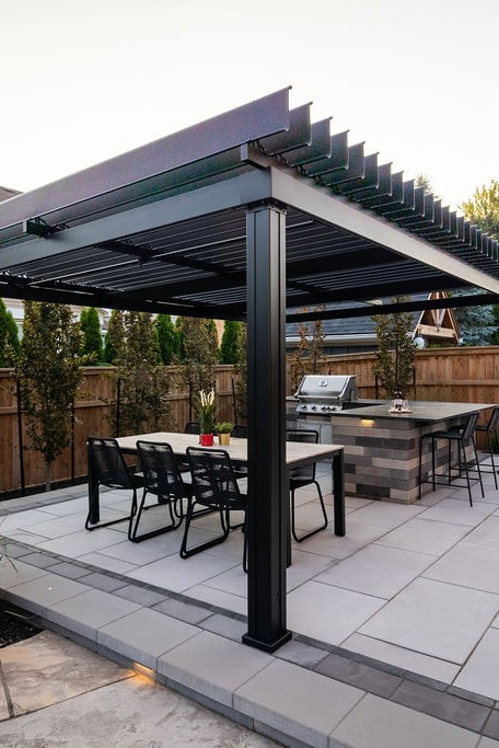 A modern outdoor patio featuring a sleek pergola, dining table, chairs, and a barbecue grill set against a wooden fence backdrop.