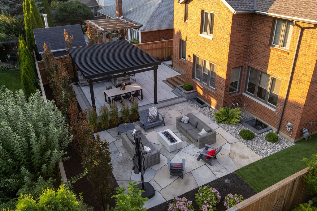 Aerial view of a modern backyard with patio furniture, fire pit, pergola, and landscaped garden, adjoining a brick house.
