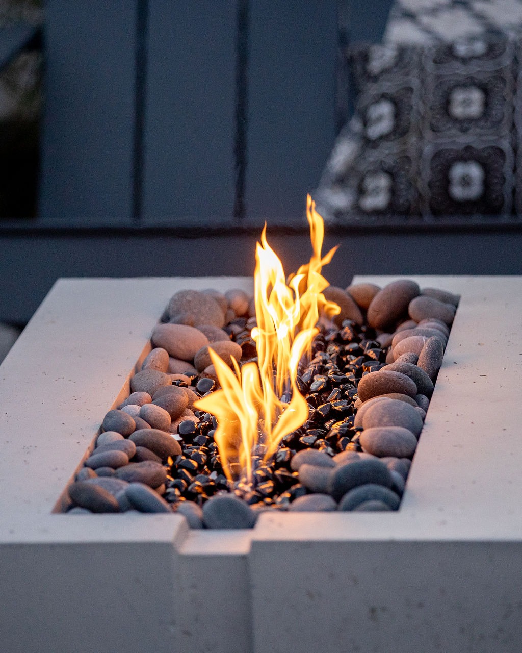 A modern rectangular fire pit with flames, surrounded by decorative stones. The background shows patterned cushions, creating a cozy outdoor setting.