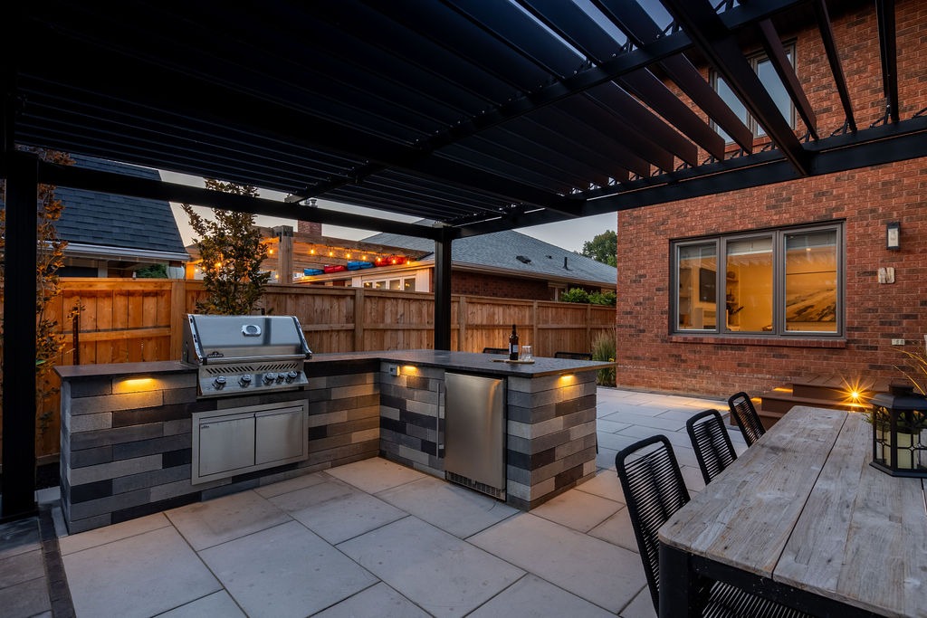Modern outdoor patio with a sleek grill, wooden table, and chairs. Twilight ambiance highlights brick architecture and warm lighting under a pergola.