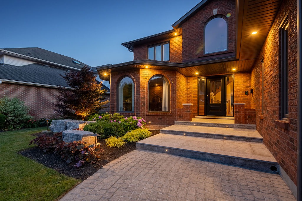 Brick house with arched windows and a neat garden. Illuminated at dusk, surrounded by well-maintained lawn and stone pathway leading to entrance.