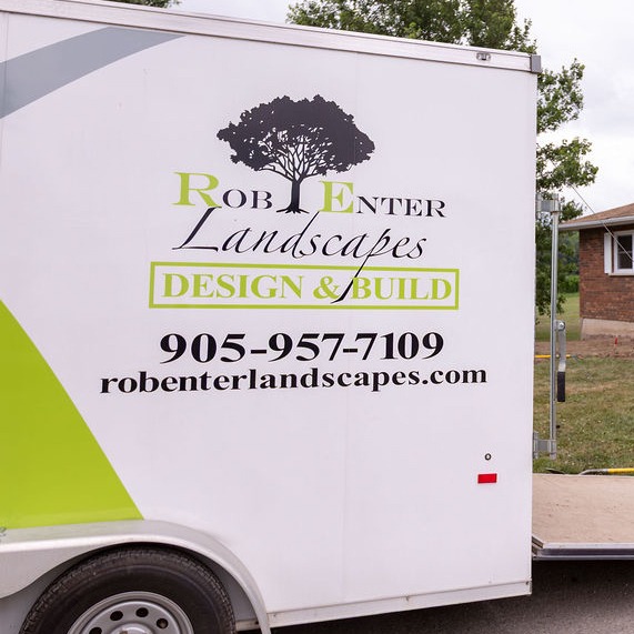White trailer with "Rob Enter Landscapes" logo and contact information, parked beside a brick house. Background features trees and grassy area.