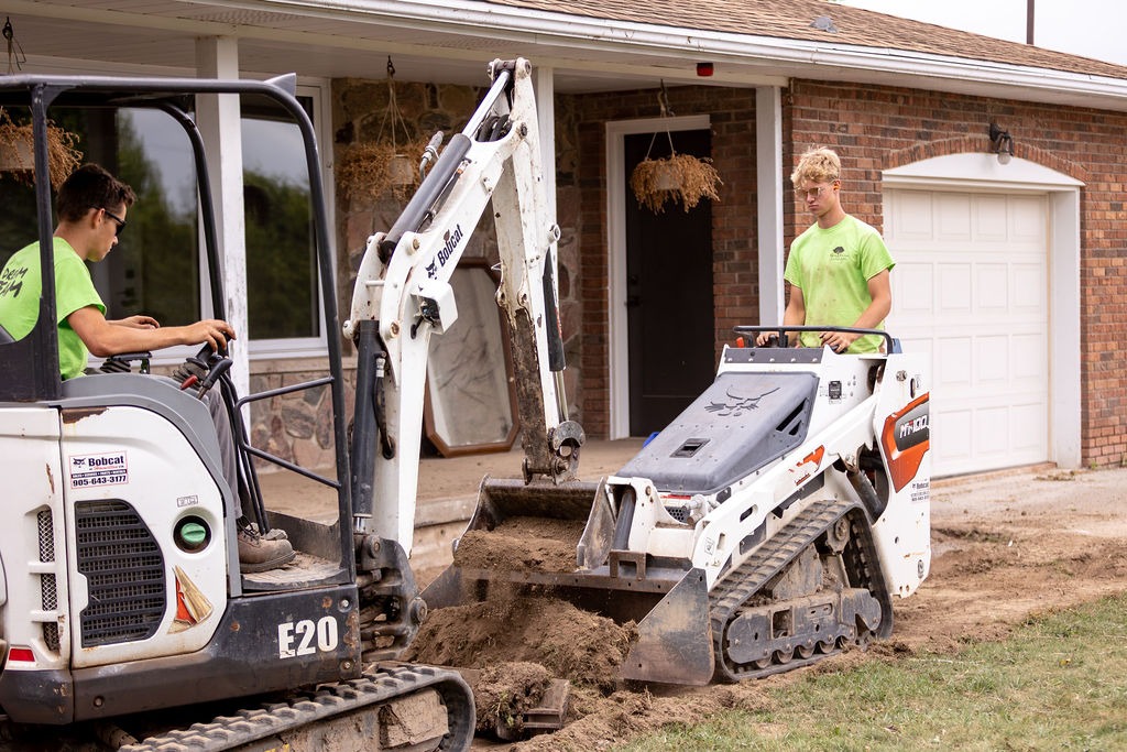 Two individuals operate compact construction equipment, digging soil in front of a brick house with hanging plants and a garage door.