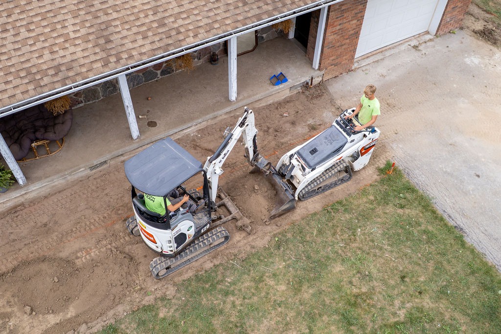 Two persons operate mini excavators on dirt beside a building. One excavation machine has a canopy, and the area is being prepared.