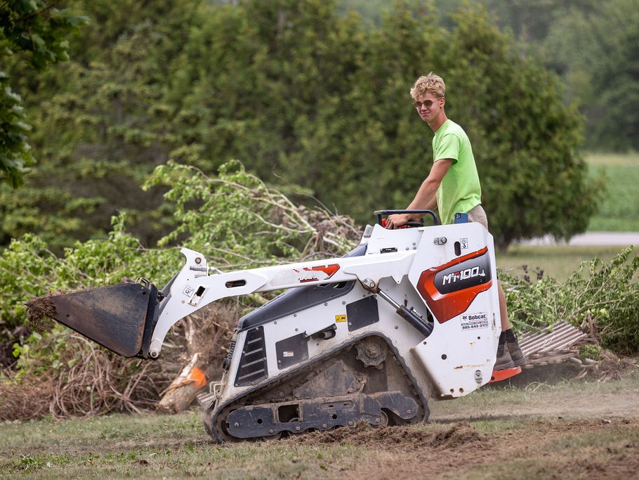 A person operates a Bobcat mini loader, clearing debris in a green landscape. Trees and shrubs are visible in the background.