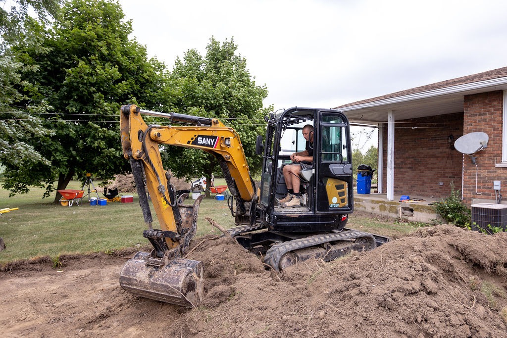 A person operates a small excavator near a brick house with a satellite dish, surrounded by trees and construction tools on grass.