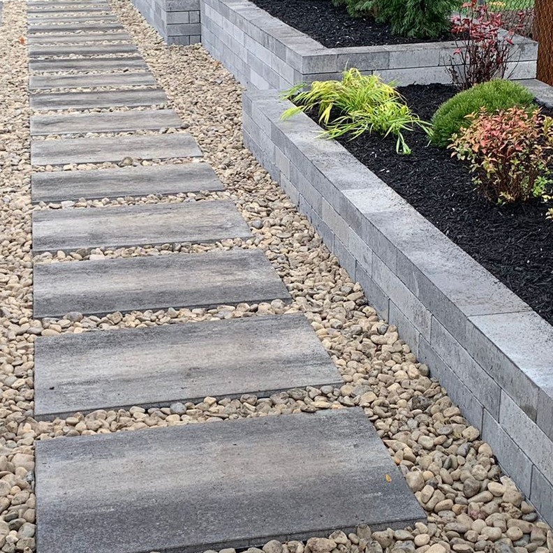 A modern garden path with rectangular stone slabs and gravel, bordered by a neatly arranged raised garden bed with various plants.