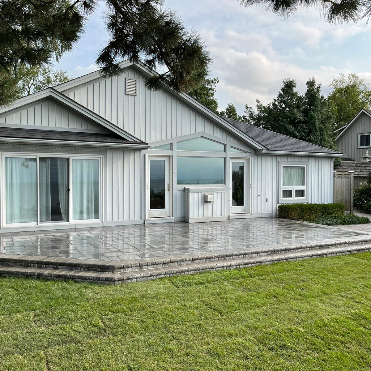A modern, single-story house with large windows and gray siding, surrounded by greenery and a manicured lawn under a cloudy sky.