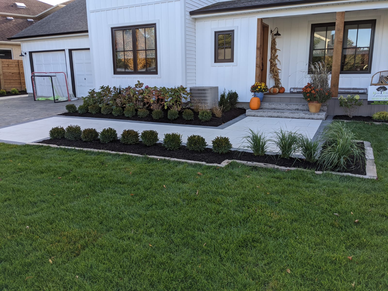Modern house with white siding, decorated porch, and neatly landscaped garden. Garage in view with a hockey net nearby.
